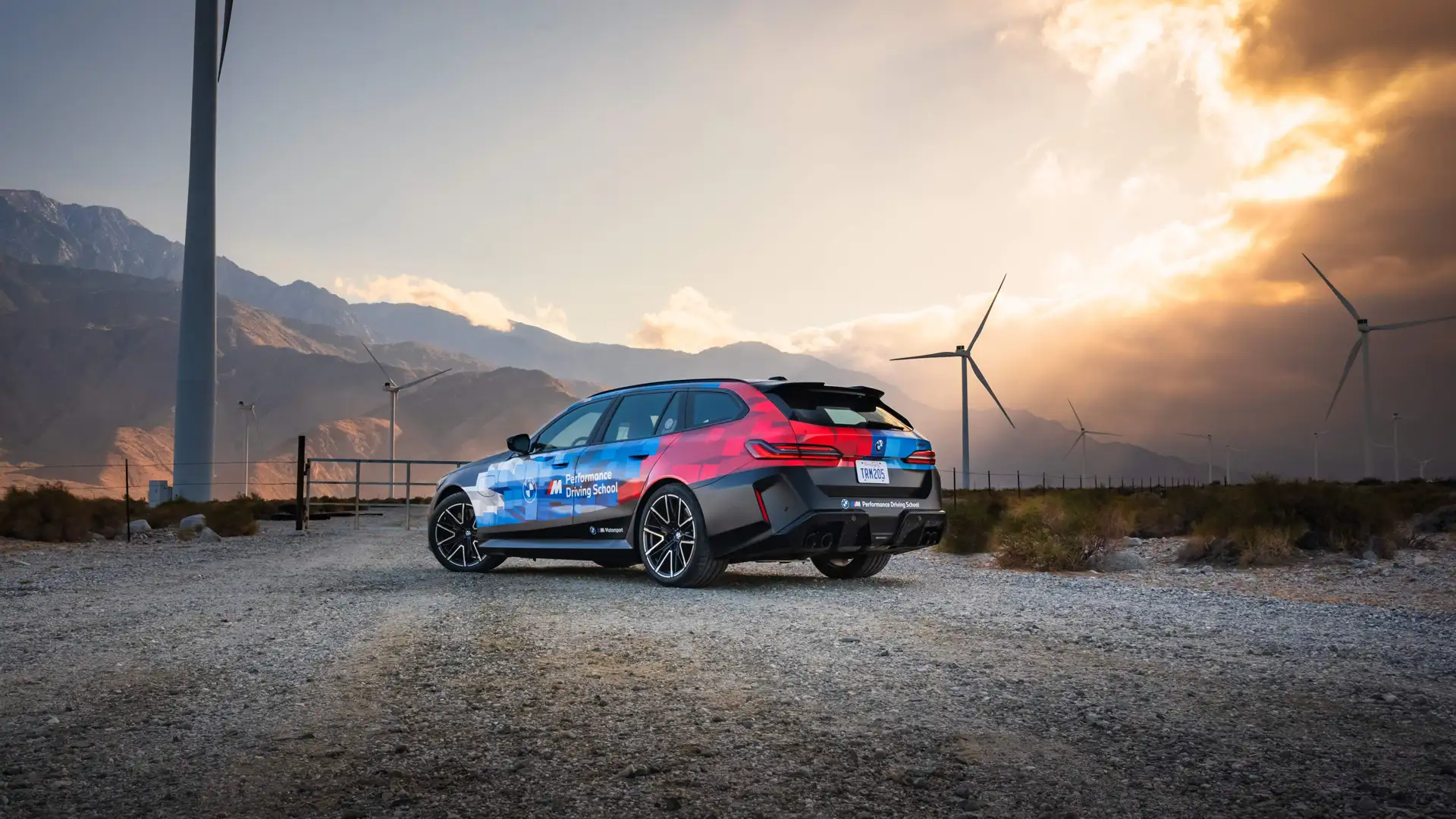 BMW M5 Touring in Performance Driving School livery among desert wind turbines at sunset