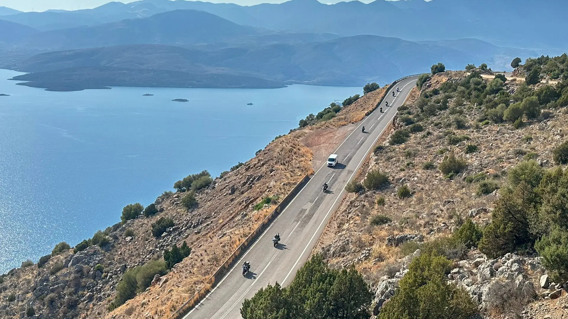 Riders strung out along a coastal mountain road above a blue bay in Greece