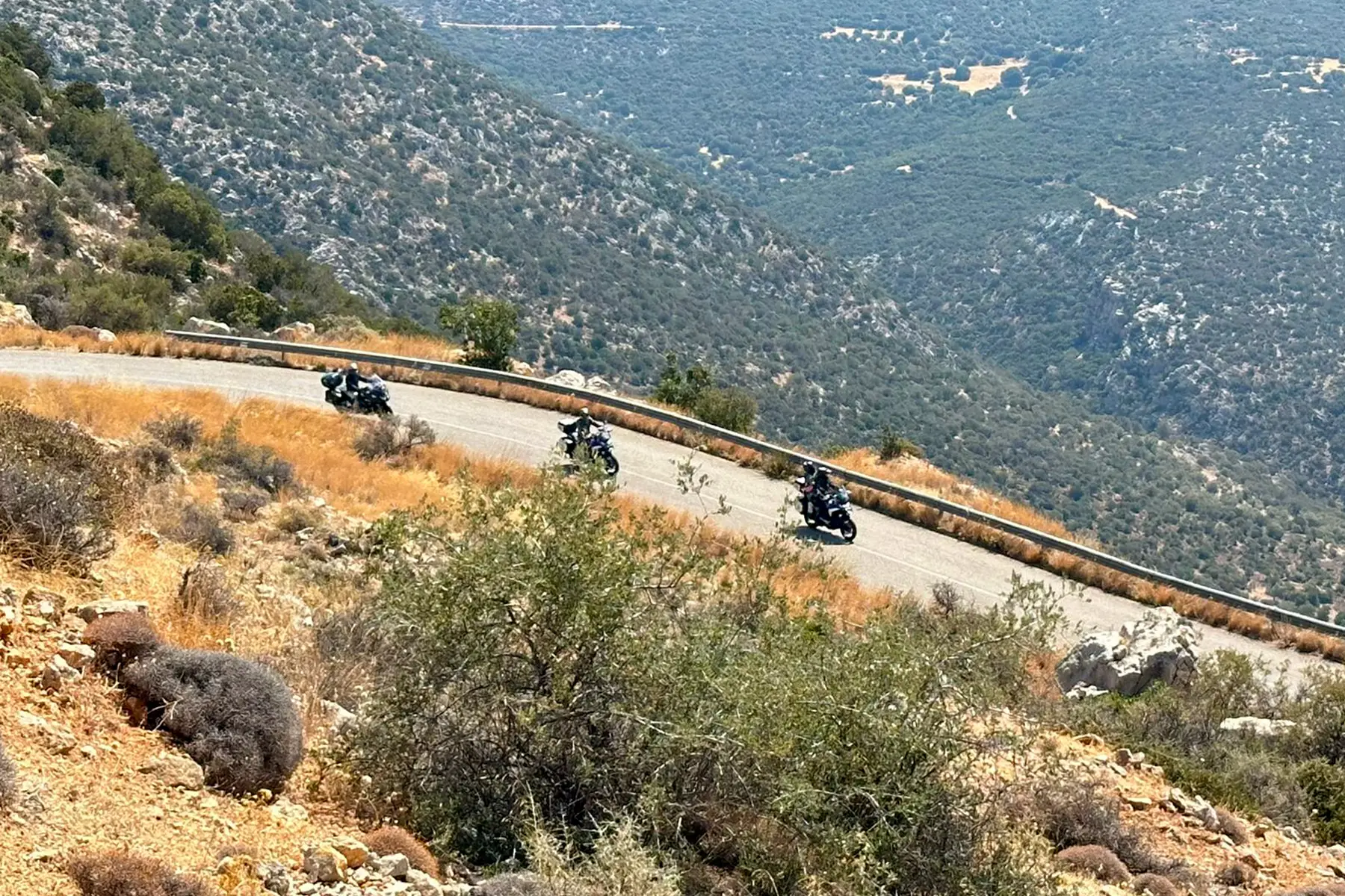 Three riders on a winding mountain road above a wooded valley